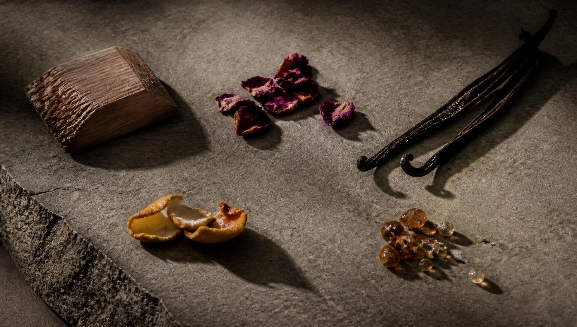 Raw fragrance materials — cedar, rose petals, vanilla pod, citrus peel and amber resin — on stone