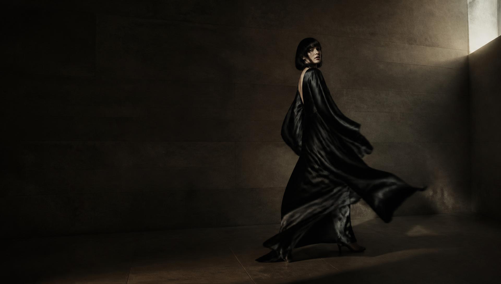 Figure in flowing dark silk moving through a bare stone room